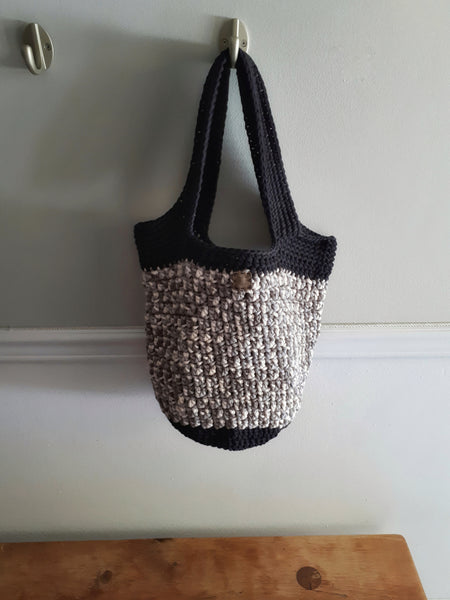 Bag hanging on wall hook over wood bench. Bag has black base and handles with olive and sand body.