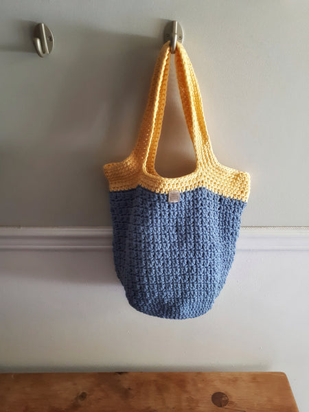 Bag hangs on wall hook over wood bench. Bag in denim blue with soft yellow trim and handles.