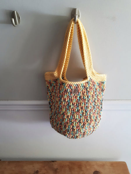 Bag hangs on wall hook over wood bench. Multi colour bag of yellow, green, and orange tropical tones. Soft yellow trim and handles.